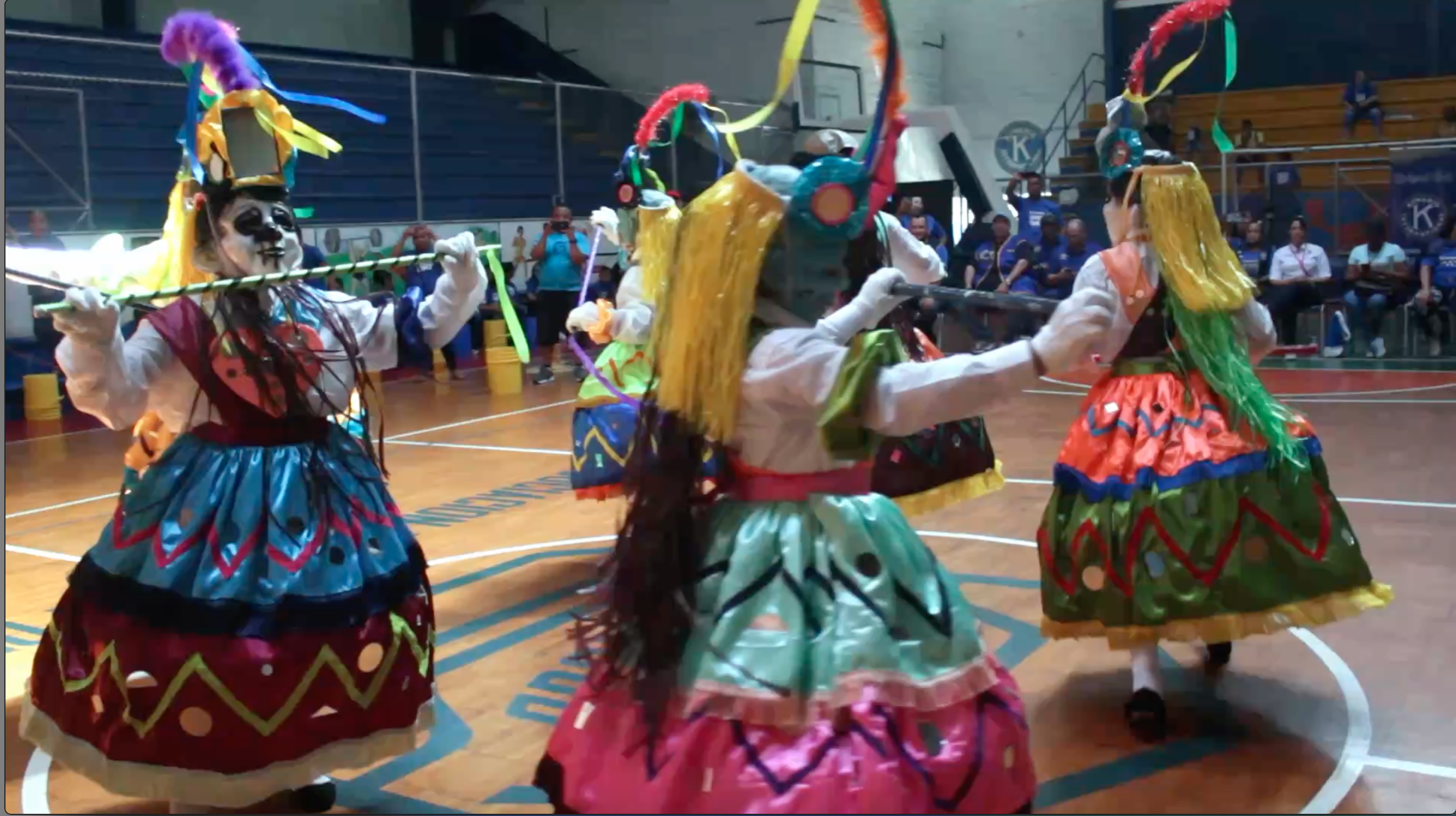 Four people are dancing in the middle of a room. They are all wearing colorful dresses, wigs and masks. In the background is an audience watching the performers.